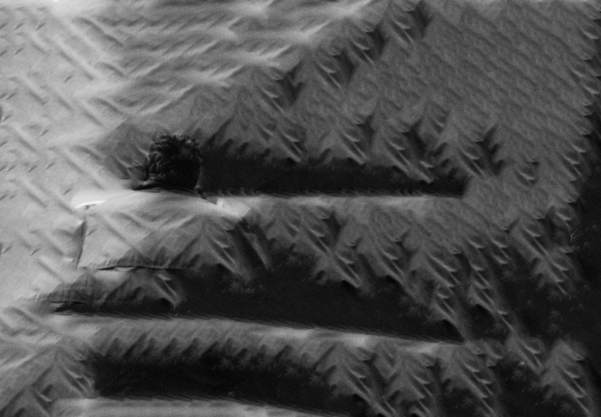 Cette photographie argentique en noir et blanc, datant de 1999, capture un moment de solitude dans un environnement urbain. On y voit une personne assise sur un banc, de dos à l’objectif, regardant la scène de la rue. L’architecture des bâtiments en face suggère un cadre européen, avec des façades caractéristiques. Des voitures sont stationnées le long de la route, et un petit kiosque ou magasin est visible. Une scène quotidienne qui évoque la vie citadine.